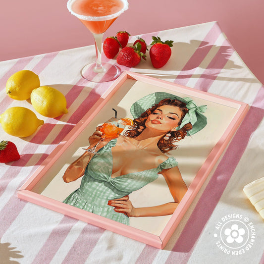 a framed illustration of a woman in a green dress holding a drink, surrounded by a pink and white striped tablecloth, lemons, strawberries, and a cocktail glass.