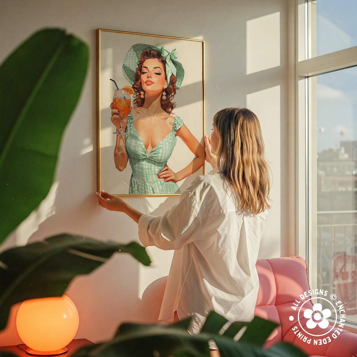 A woman hangs a framed vintage-style illustration of a woman in a green dress and hat on a wall in a room with a pink chair and a plant.