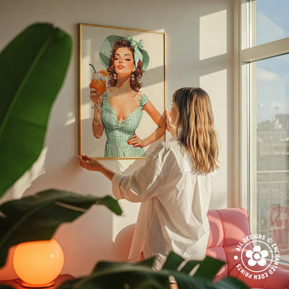 A woman hangs a framed vintage-style illustration of a woman in a green dress and hat on a wall in a room with a pink chair and a plant.