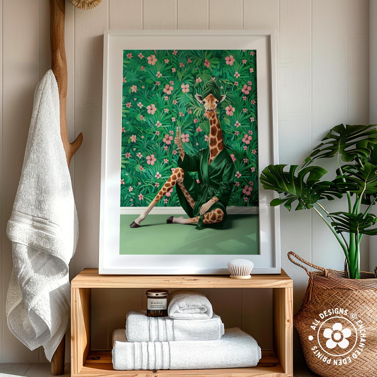 a framed artwork of a giraffe sitting on a green surface, surrounded by a floral background. The artwork is displayed on a wooden shelf, with a towel hanging nearby and a potted plant in the background.