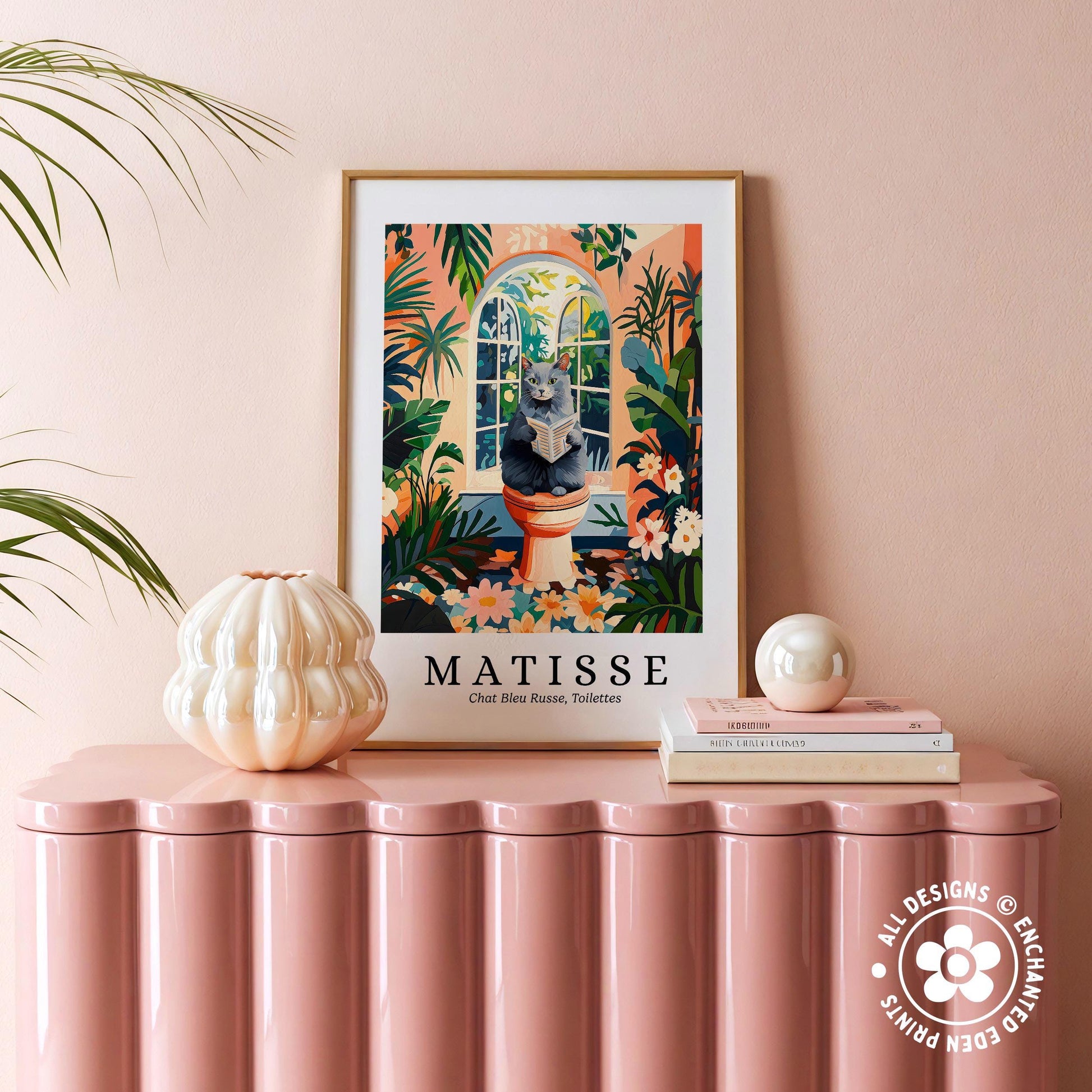 a framed poster of a cat sitting in a window, surrounded by tropical plants, placed on a pink table.