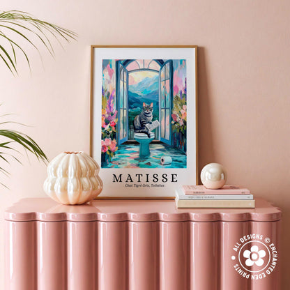 a framed painting of a cat sitting on a toilet, surrounded by a colorful and whimsical scene, placed on a pink table.