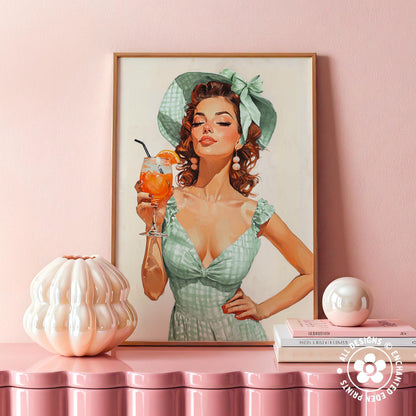 a framed painting of a woman in a green dress holding a drink, with a pink wall in the background. There is also a small white vase and a stack of books on a pink surface.