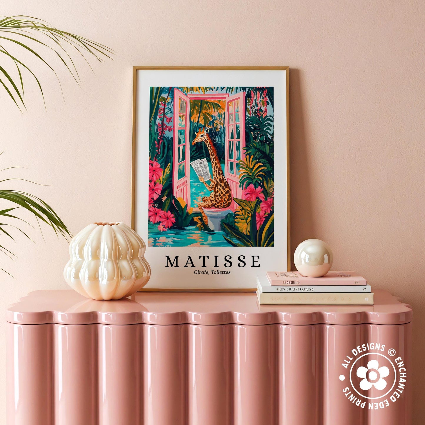 a pink table with a framed poster of a giraffe in a tropical setting, surrounded by flowers and plants, placed on top of a stack of books.