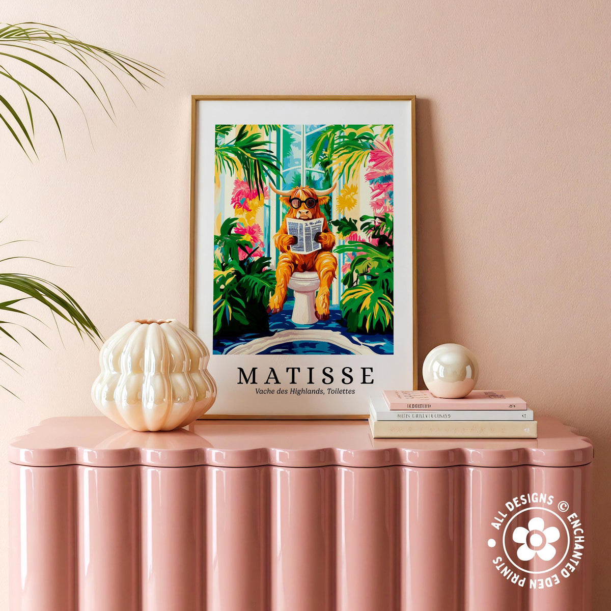 a framed poster of a cat sitting on a toilet, surrounded by tropical plants, with the word "MATISSÉ" prominently displayed. The poster is placed on a pink surface, and there are some decorative items, such as a white vase and a stack of books, nearby.