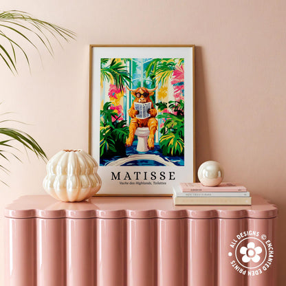 a framed poster of a cat sitting on a toilet, surrounded by tropical plants, with the word "MATISSÉ" prominently displayed. The poster is placed on a pink surface, and there are some decorative items, such as a white vase and a stack of books, nearby.
