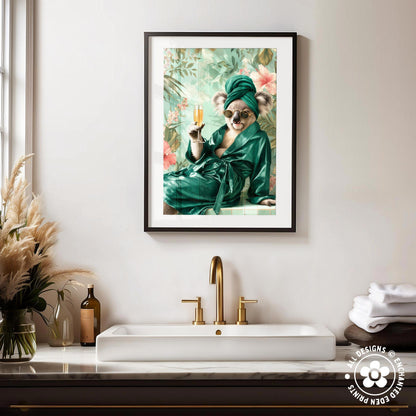 a framed picture of a koala wearing a green robe and holding a glass of champagne, sitting on a white marble countertop in a bathroom setting.