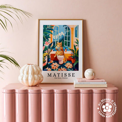 a framed poster of a cat sitting on a toilet, surrounded by tropical plants, placed on a pink table.
