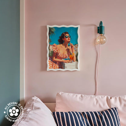 a framed vintage-style photograph of a woman in a red and white dress, sitting on a couch and holding a drink. The photograph is hanging on a pink wall above a striped pillow.