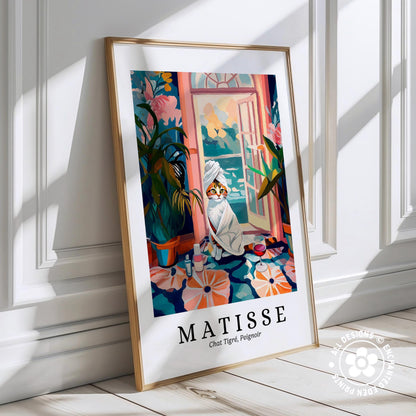 A framed poster depicting a cat wrapped in a towel, sitting on a windowsill with a potted plant and flowers in the background, against a white wall.