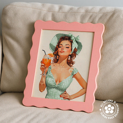 A framed painting of a woman in a green dress holding a glass of orange juice, with a scalloped pink frame.