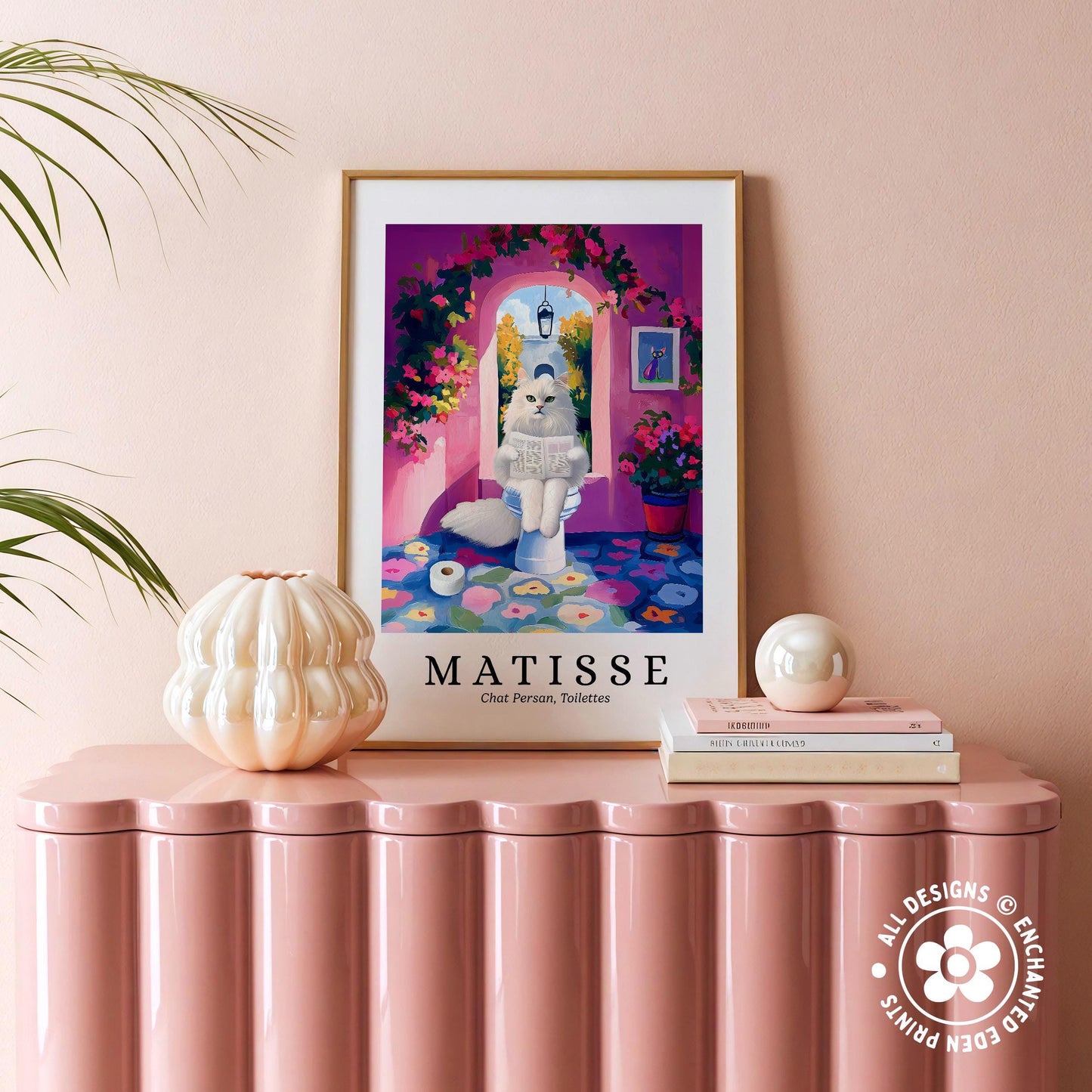 a framed poster of a whimsical painting of a cat sitting on a toilet, surrounded by a colorful and playful scene. The poster is displayed on a pink table, with a white vase, a stack of books, and a small white ball nearby.