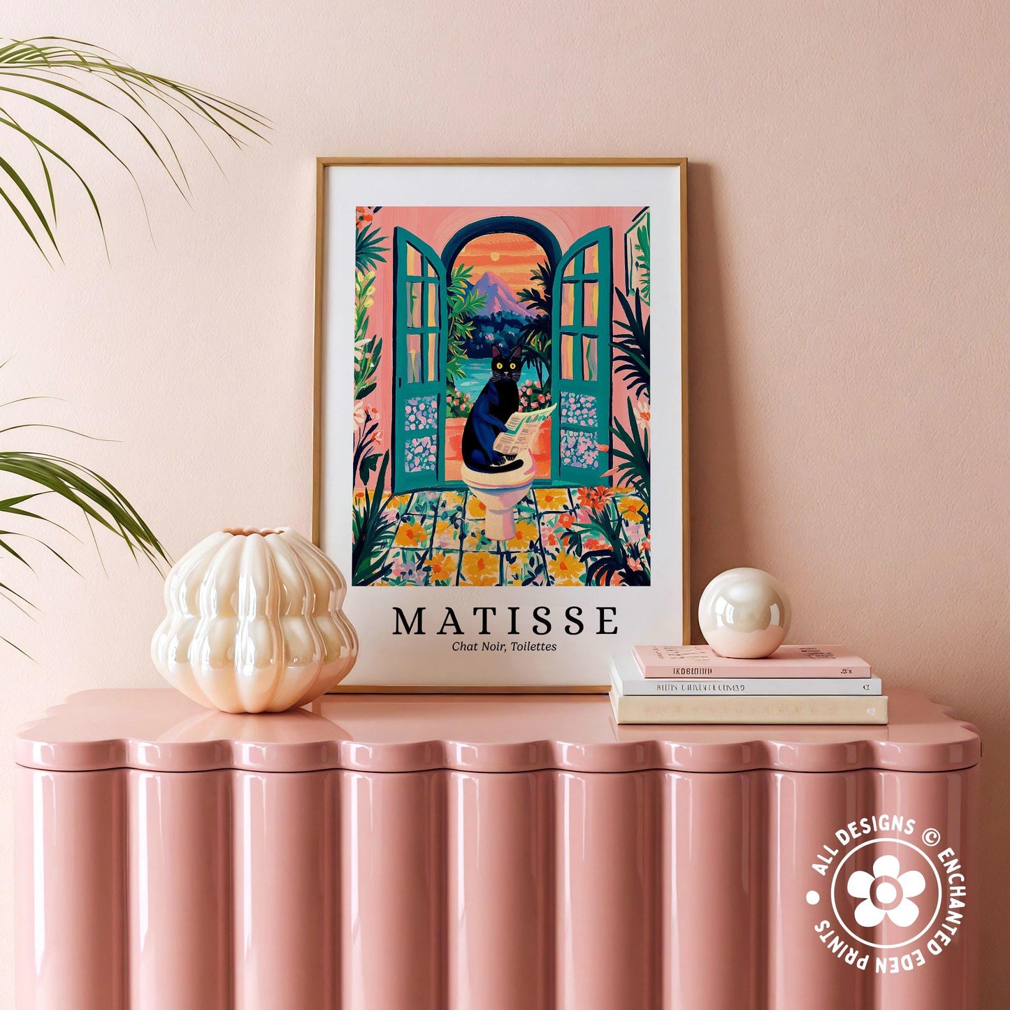 a pink table with a framed poster of a colorful illustration of a cat sitting in an open window, surrounded by tropical plants. The poster is titled "MATISSE" and is framed in a white frame with a gold border. The table also has a white vase and a stack of books.