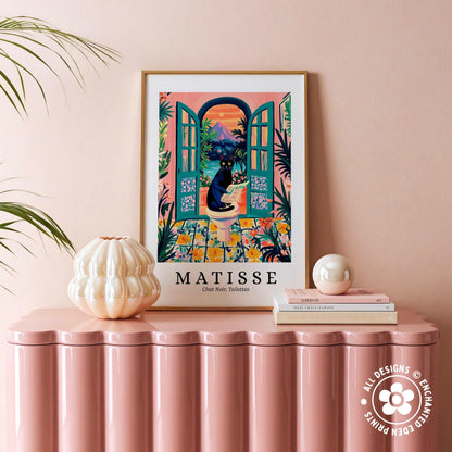 a pink table with a framed poster of a colorful illustration of a cat sitting in an open window, surrounded by tropical plants. The poster is titled "MATISSE" and is framed in a white frame with a gold border. The table also has a white vase and a stack of books.