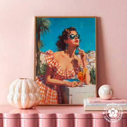 a framed painting of a woman in a red and white checkered dress, holding a glass of orange juice and looking up at the sky. The painting is displayed on a pink shelf, with a white vase and a stack of books placed next to it.
