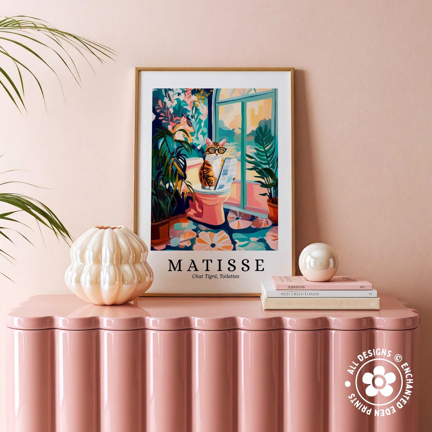 a framed poster of a cat sitting on a pink stool, with a window and plants in the background, placed on a pink table.