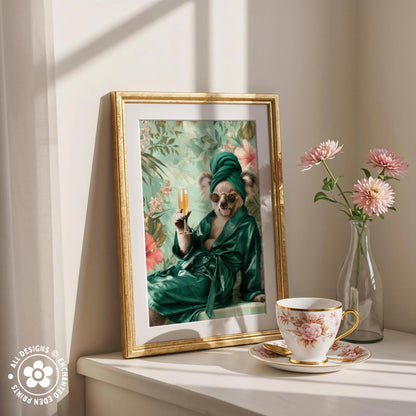 A framed painting of a koala wearing a green robe and holding a champagne glass is displayed on a white shelf next to a vase of pink flowers and a teacup.