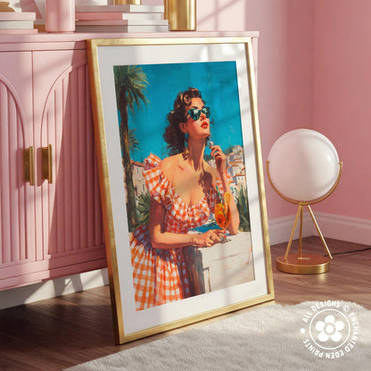 A framed vintage-style painting of a woman in a red and white checkered dress, wearing sunglasses and holding a drink, is displayed on a pink cabinet in a room with a white lamp and a pink wall.